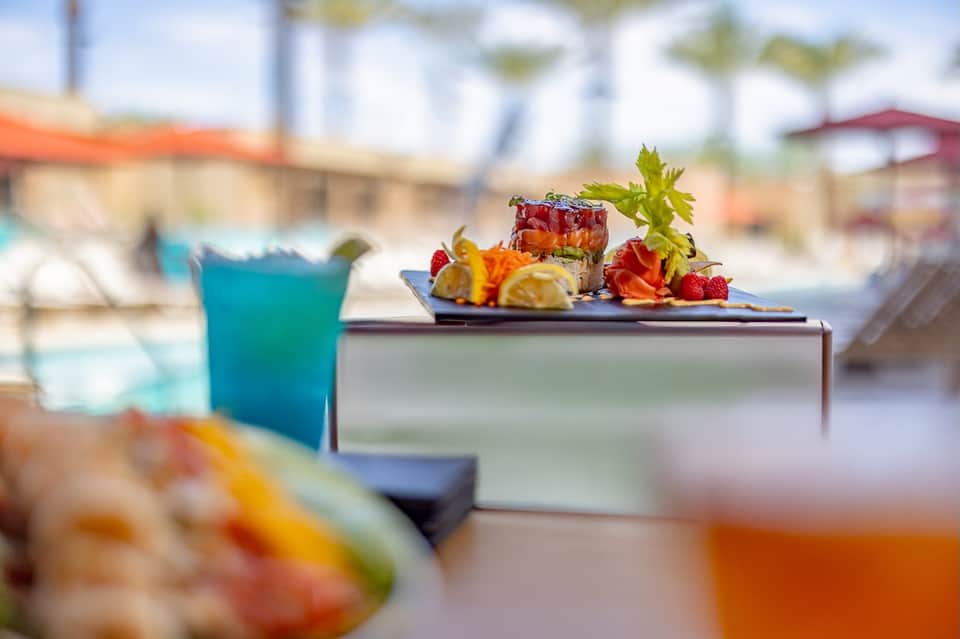 Colorful tropical cocktail with fruit garnish on poolside table, blurred beach and palm trees in background