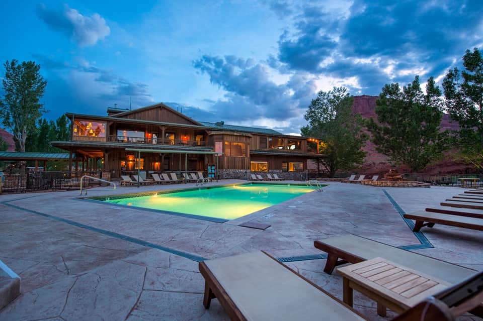 Illuminated resort pool at dusk with rustic lodge building, mountains, and lounge chairs in mountain setting