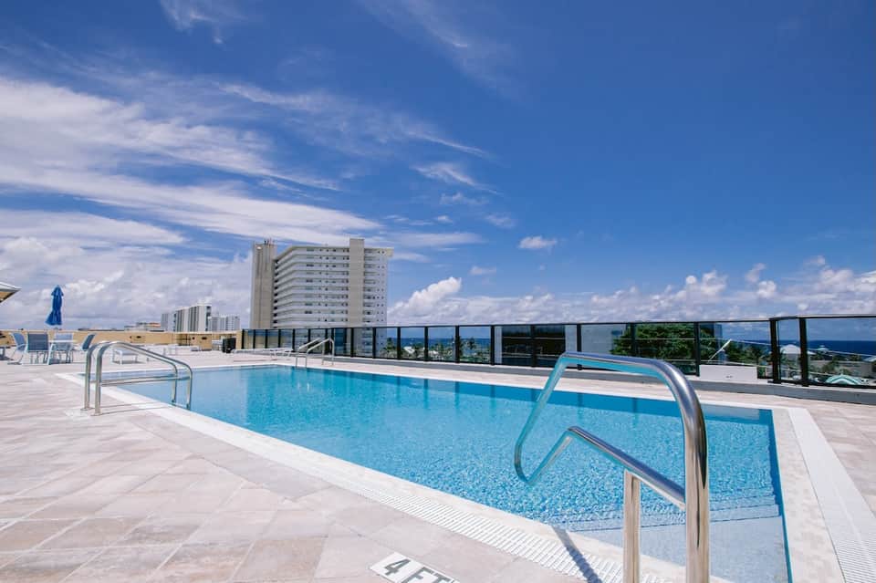 Rooftop swimming pool with metal railings, city buildings, and blue sky with clouds
