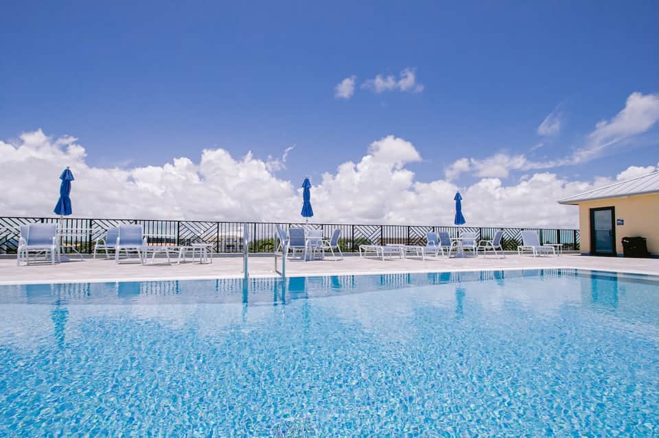 Resort pool with white loungers, blue umbrellas, and ocean views beyond railing
