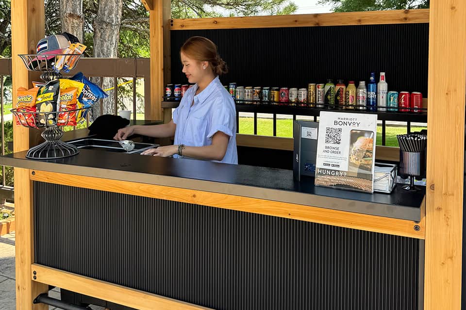 Outdoor kiosk with wooden frame, black metal counter, beverage display, snack basket, and natural tree surroundings