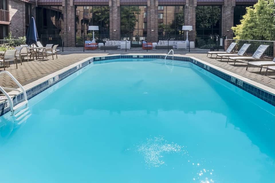 Outdoor swimming pool with turquoise water, lounge chairs, blue umbrella, and brick building backdrop.