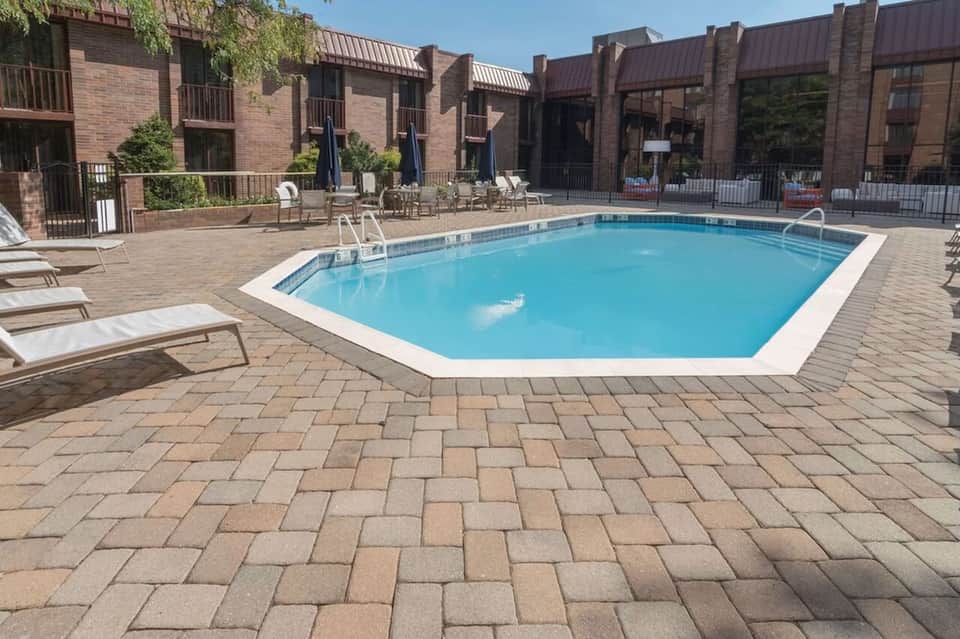 Resort pool with brick buildings, loungers, umbrellas, fencing, and patio area in sunny weather