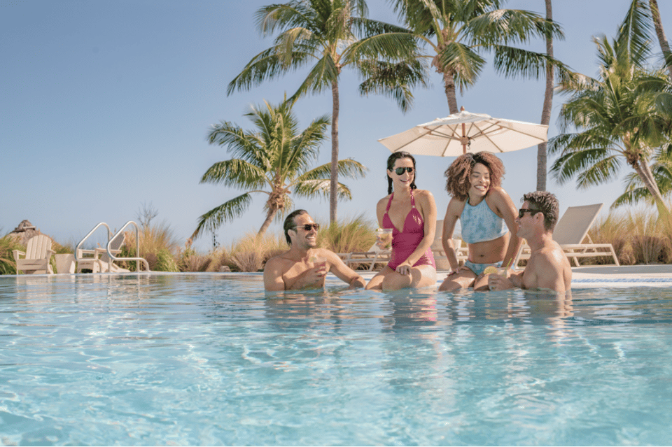 Family swimming in resort pool with palm trees, white umbrellas, and tropical setting