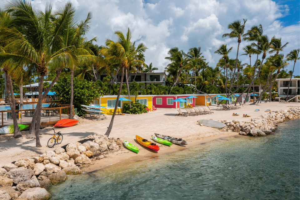 Colorful beach huts and palm trees along sandy shore with water sports equipment and resort buildings