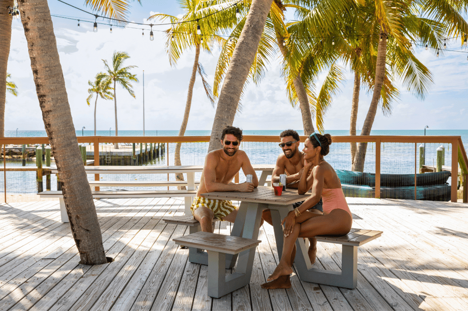 Three people enjoying drinks on a wooden deck overlooking the ocean with palm trees