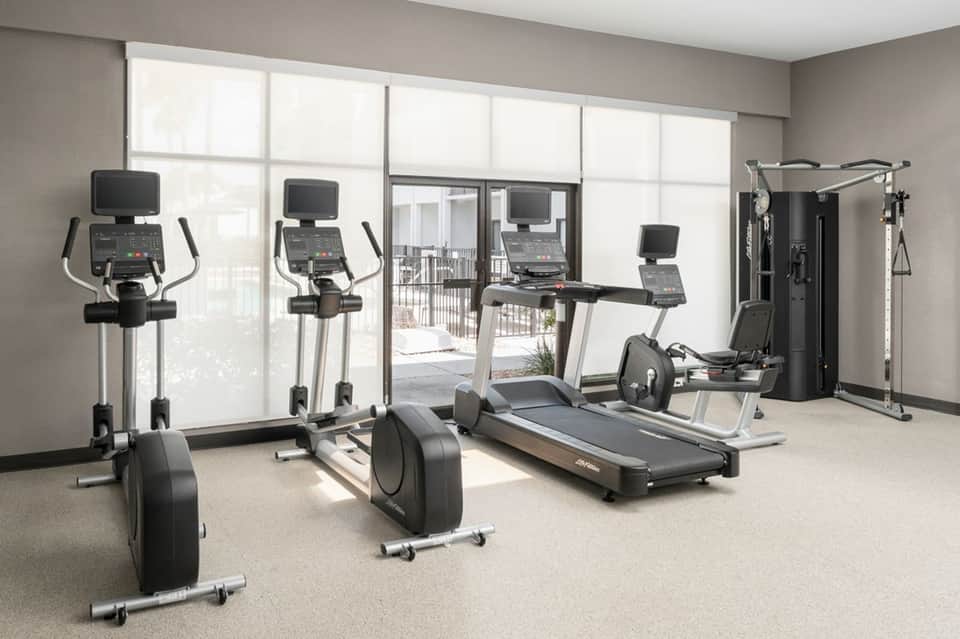 Bright fitness center with cardio equipment, treadmills, ellipticals, and large windows overlooking a balcony