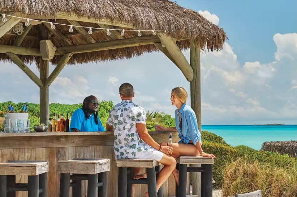 Tropical beachside tiki bar with thatched roof, turquoise ocean view, bartender serving couple, lush vegetation