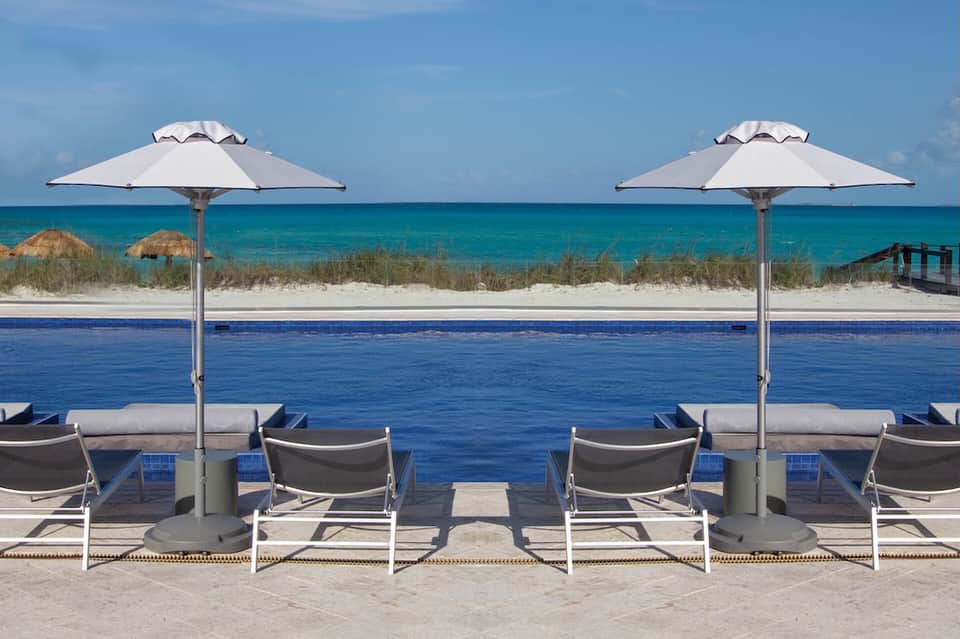 Beach resort pool overlooking turquoise ocean with white umbrellas and sandy beach