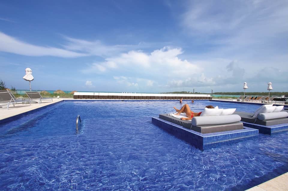 Rectangular pool with floating loungers, ocean and sky view, white umbrellas, and woman relaxing on white cushioned seating