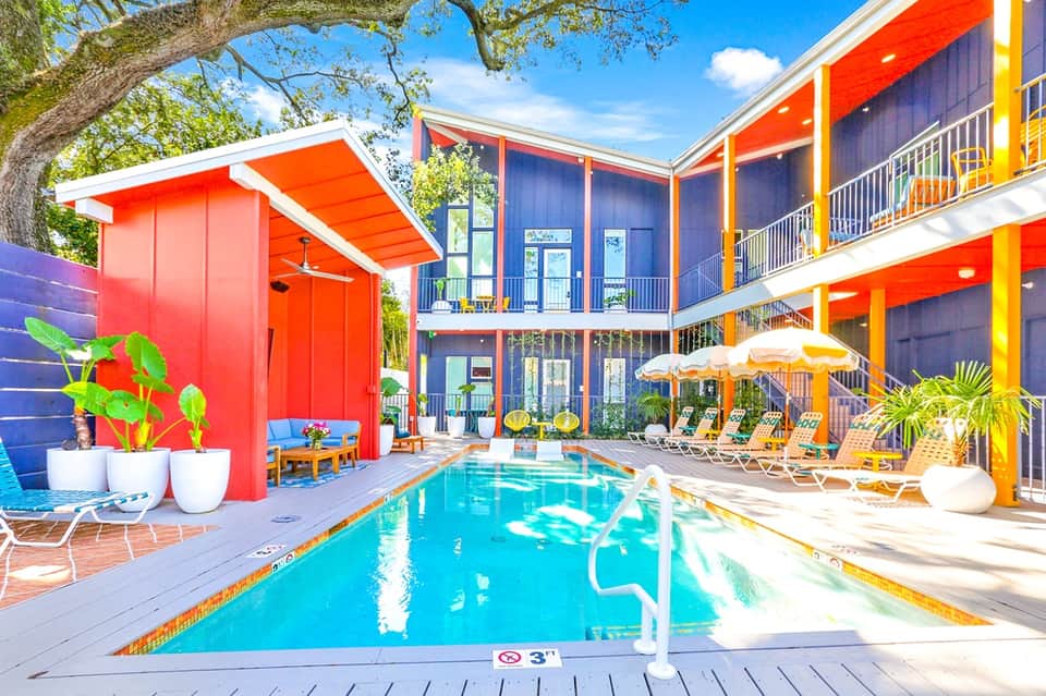 Colorful mid-century resort with turquoise pool, orange and blue buildings, lounge chairs, and palm trees