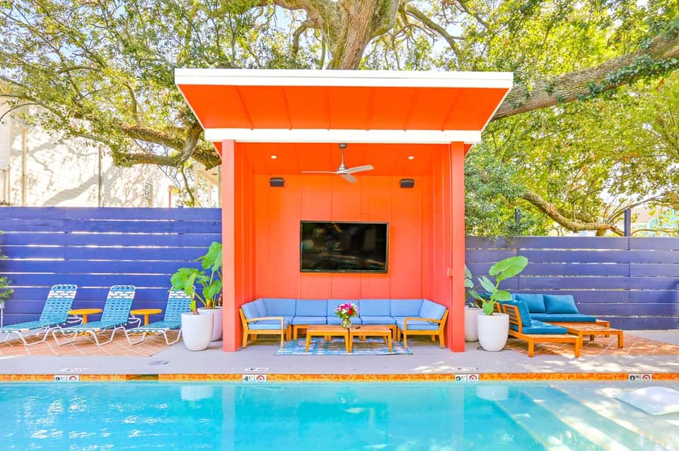 Poolside cabana with orange exterior, blue cushioned seating, and TV under mature oak trees
