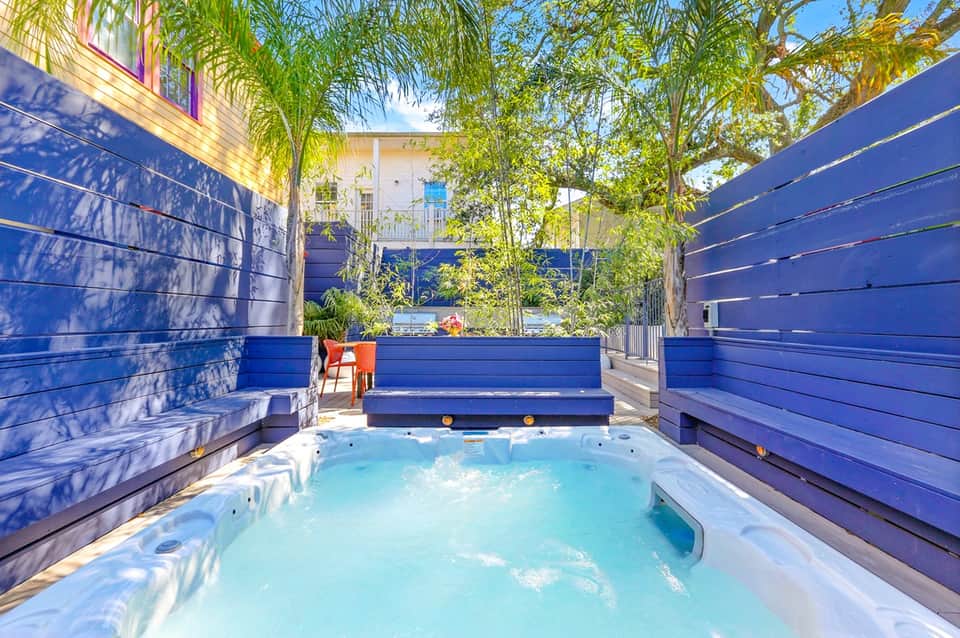 Modern hot tub enclosed by blue wooden walls with palm trees and building visible overhead