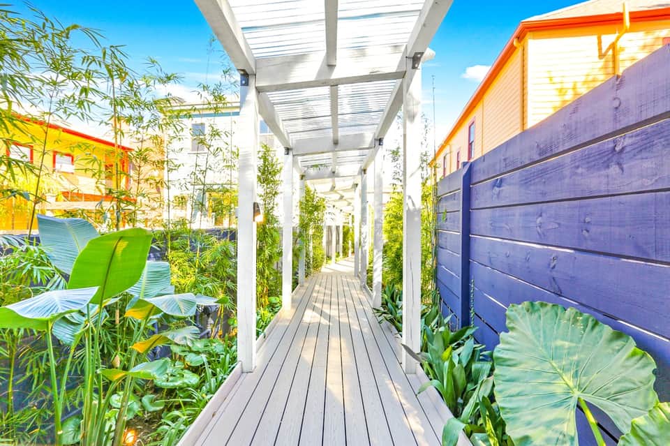 Long narrow garden pathway with white pergola, climbing vines, colorful exterior walls, and natural sunlight filtering through