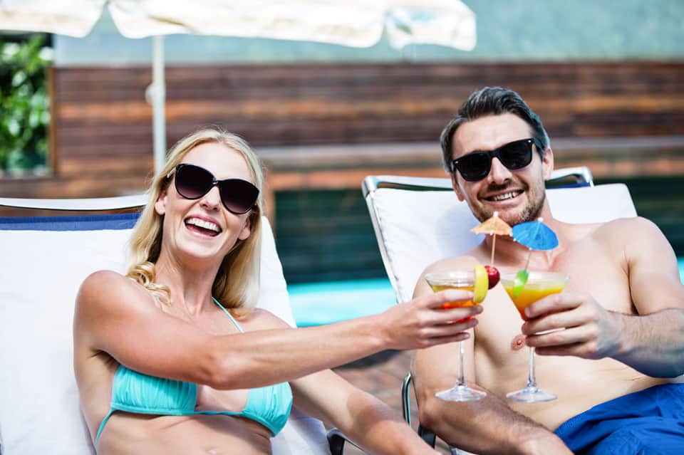 Couple in swimwear toasting with tropical cocktails by poolside, both wearing sunglasses