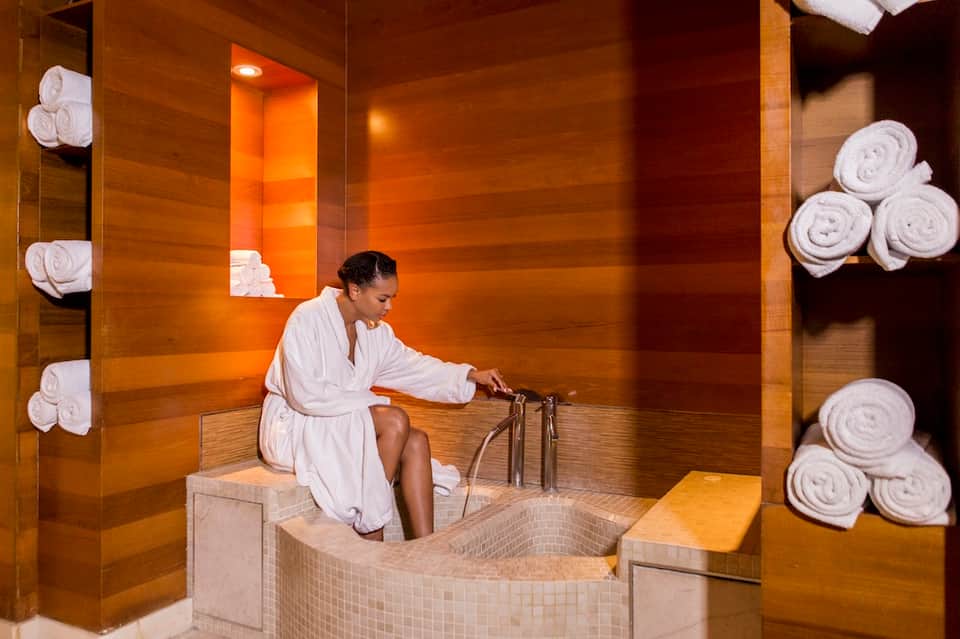 Spa steam room with wood paneling, woman in white robe using hydrotherapy, rolled towels on shelves
