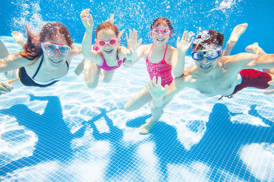 Children wearing goggles swimming underwater in a bright blue pool, waving at the camera