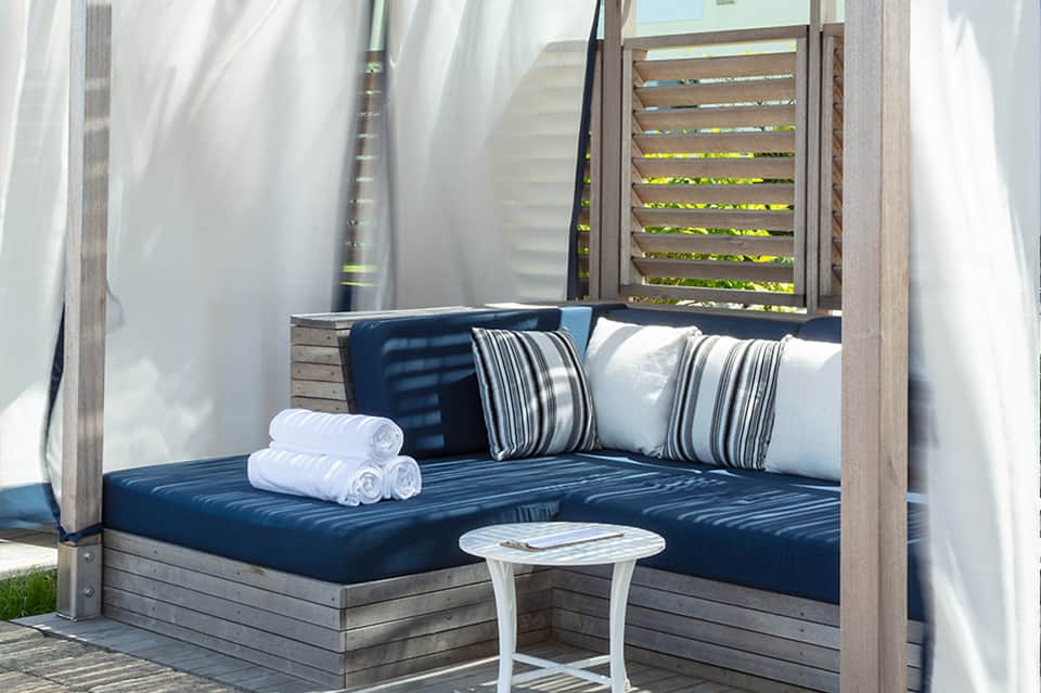 Outdoor daybed with blue cushions, striped pillows, white curtains, and shaded area