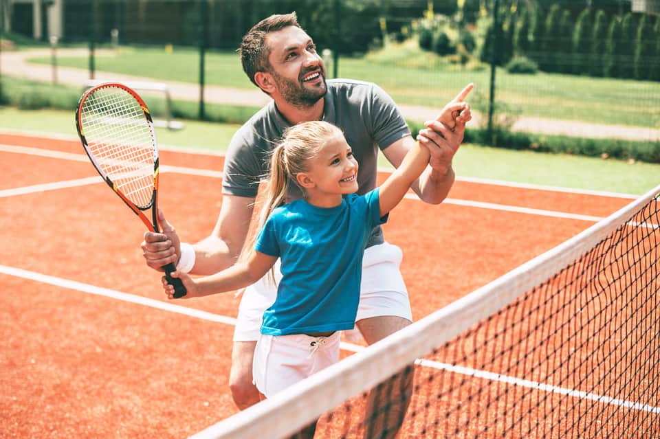 Adult teaching child tennis on red clay court with green fence and trees