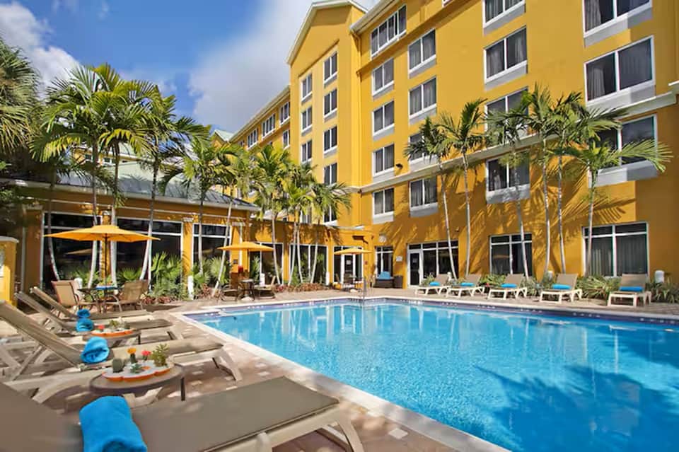 Resort outdoor pool area with lounge chairs, palm trees, yellow building facade, and blue umbrellas