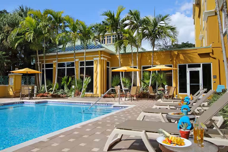 Resort pool area with yellow cabana, palm trees, lounge chairs, and tropical poolside dining setup