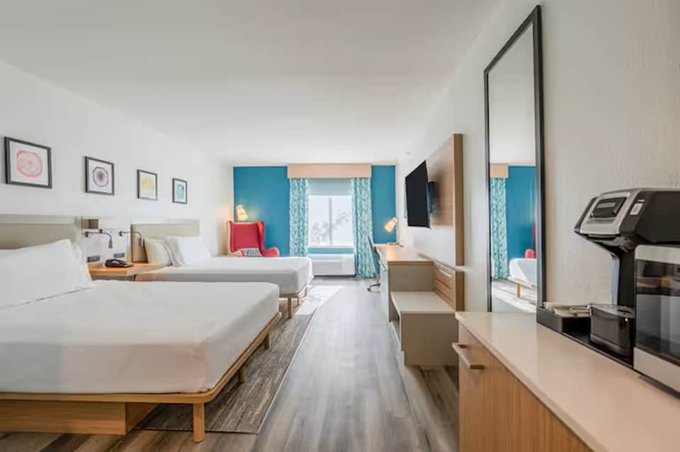 Bright hotel room with two beds, turquoise accents, wooden furniture, and treadmill