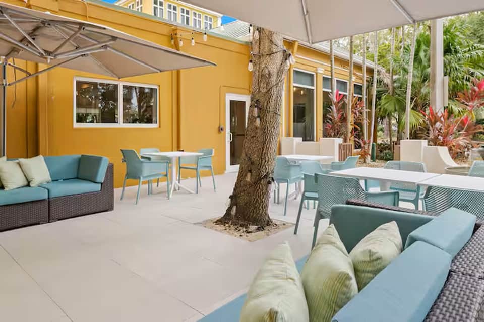 Outdoor patio with yellow building, blue lounge furniture, tree, umbrellas, and white seating