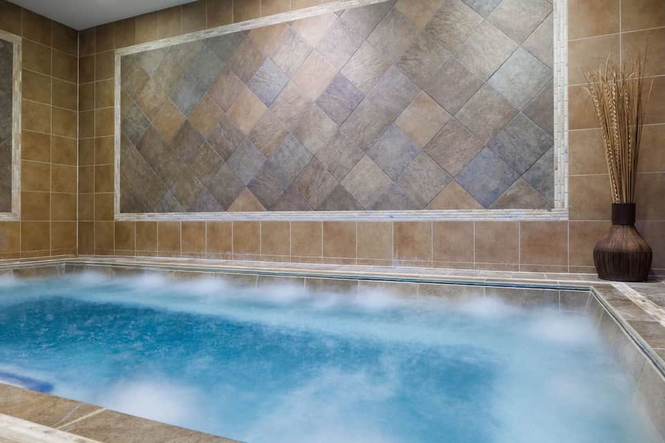 Indoor hot tub with decorative tile wall, beige surround, and spa ambiance