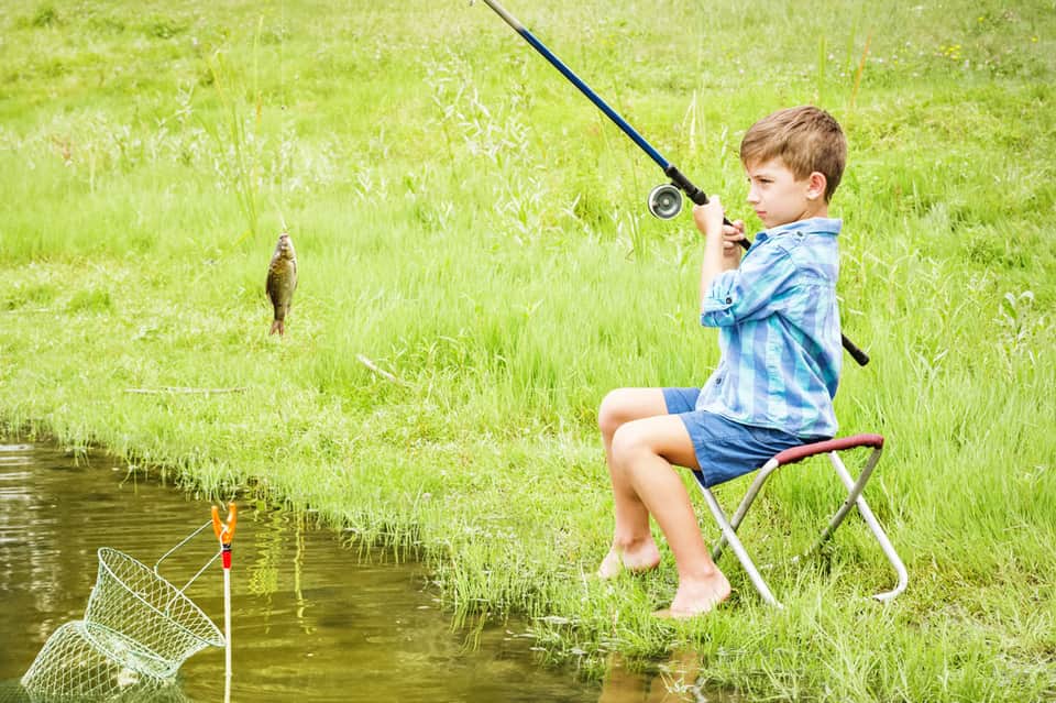 Young boy fishing from chair beside pond with green grass and fishing net