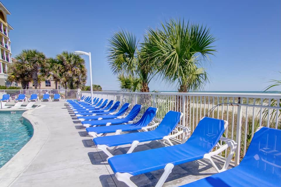 Beachfront pool with blue lounge chairs, ocean view, palm trees, protective railings, and coastal residential building