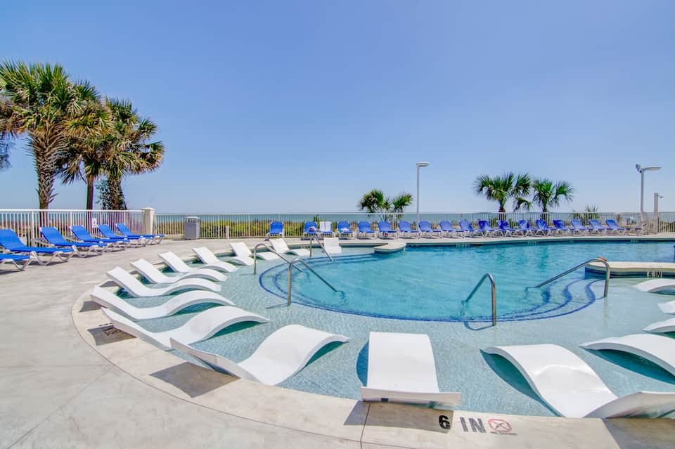 Curved swimming pool with white lounge chairs, blue lounge chairs in background, palm trees, and ocean view over railing