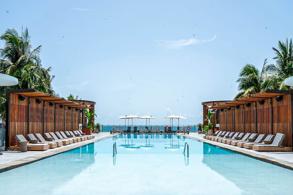 Luxury resort pool with wooden cabanas, striped loungers, palm trees, and ocean horizon view