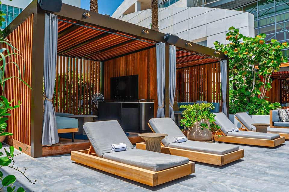 Luxury cabanas with wooden lounge chairs, pergolas, curtains, plants, and modern architecture