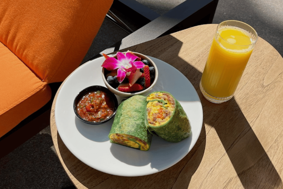 Breakfast plate with green wrap, fruit bowl, salsa, fresh juice, and orange pillow in sunlight