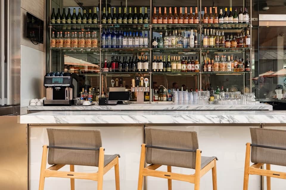 Modern hotel bar with marble counter, wooden stools, extensive bottle display behind glass shelves