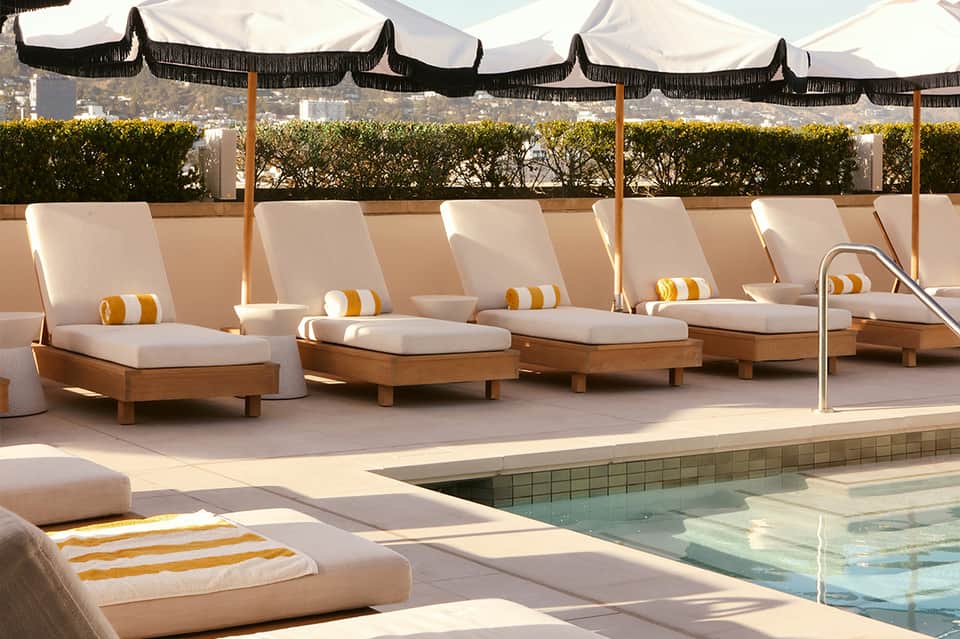 Luxury poolside with cream loungers under striped awnings, white umbrellas, hedges, and mountain view