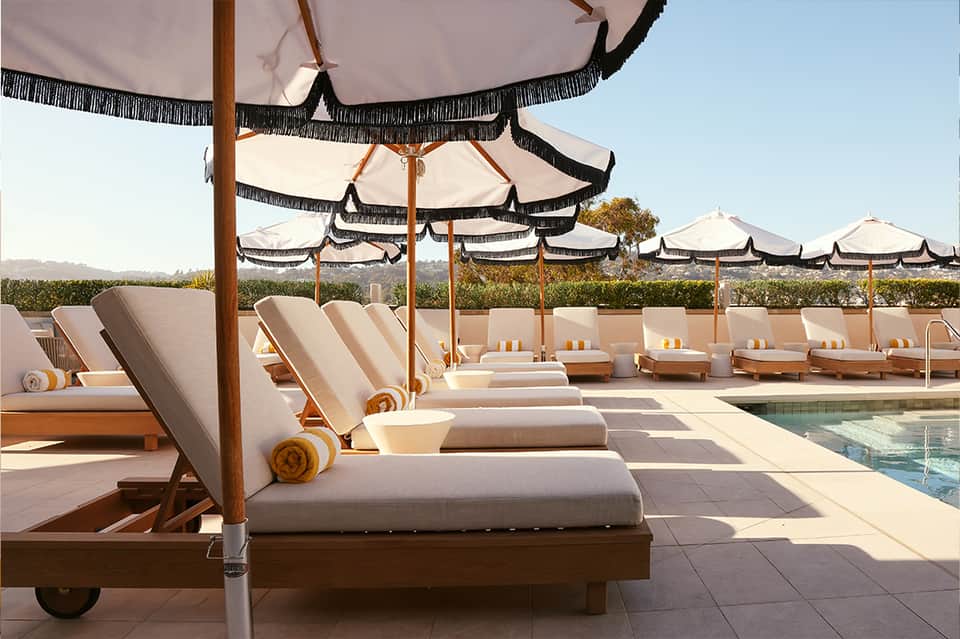 Row of lounge chairs with cream cushions under umbrellas with black fringe trim beside a pool