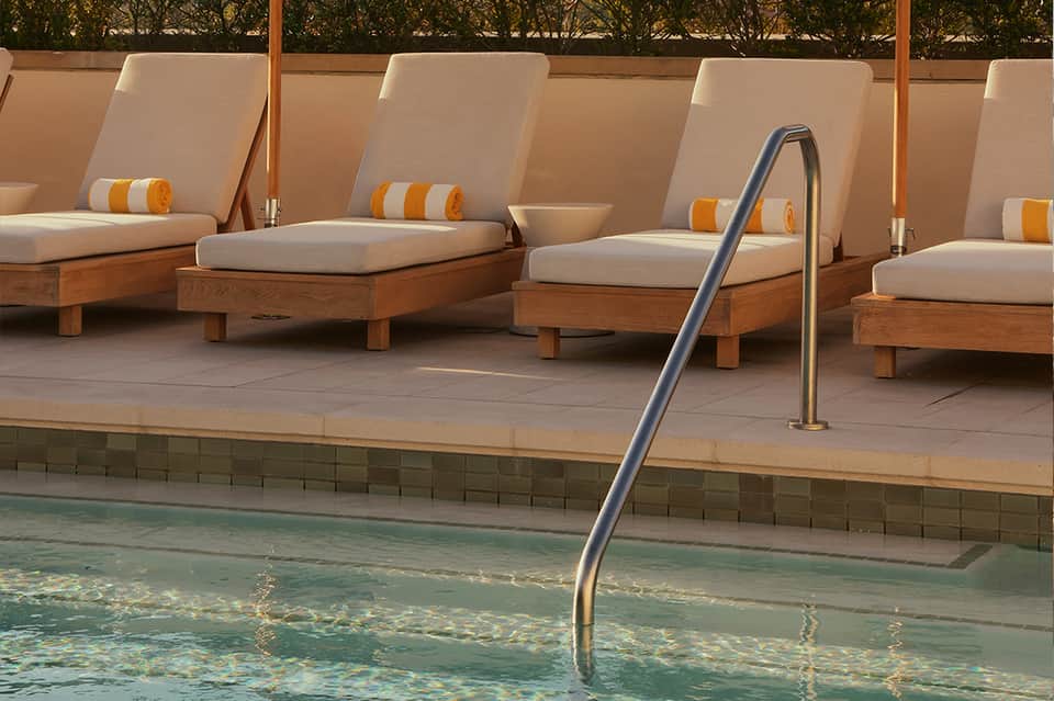 Pool deck with wooden lounge chairs, cream cushions, and metal railing beside turquoise water