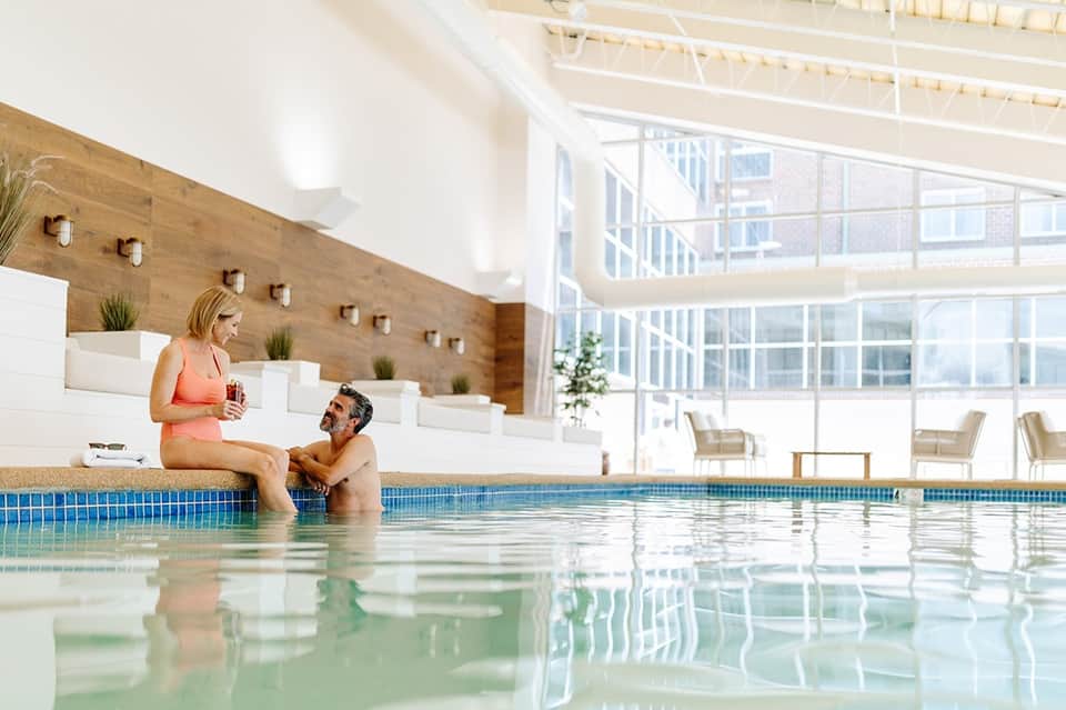 Indoor pool area with couple at pool edge, wood-paneled walls, skylights, and lounge seating