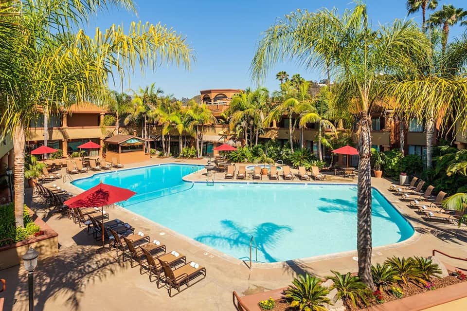 Resort pool area with multiple swimming pools, red umbrellas, palm trees, and lounge chairs