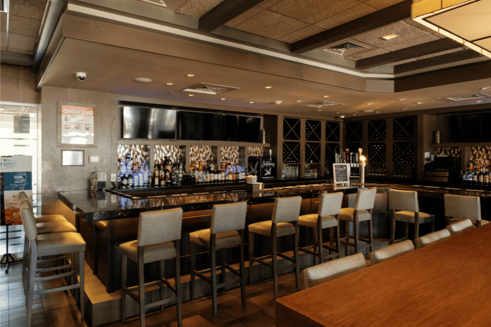 Hotel bar interior with dark wood counter, wine storage, bar seating, and warm accent lighting