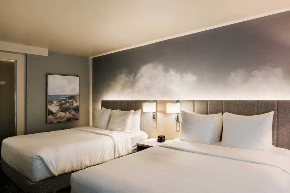 Hotel guest room with two queen beds, gray upholstered headboards, white linens, and beach artwork