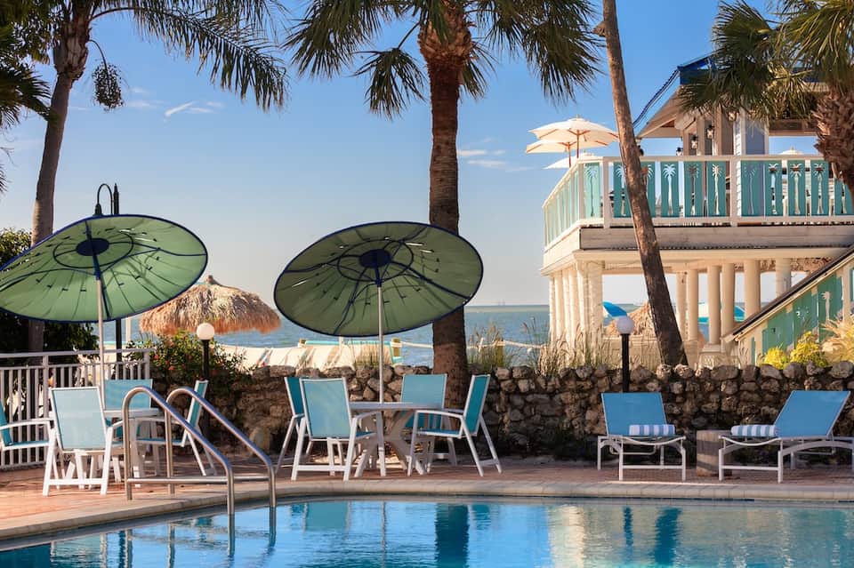 Beachfront pool area with green umbrellas, loungers, and ocean view through palm trees