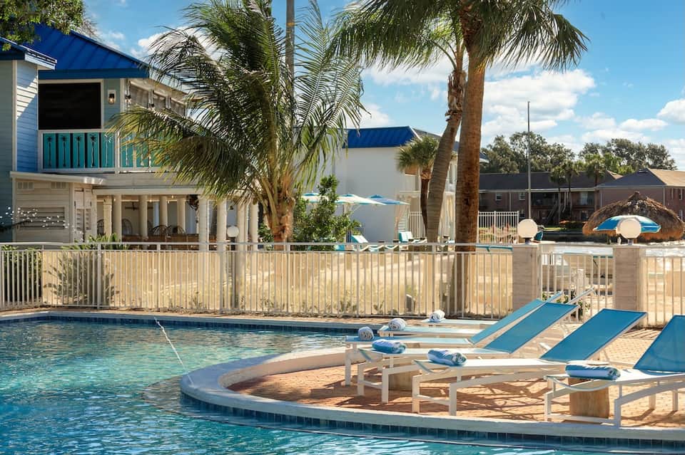 Beachfront resort pool with blue lounge chairs, spa area, palm trees, and elevated homes