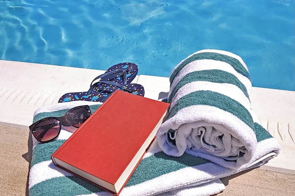 Poolside relaxation items including striped towel, sunglasses, flip-flops, and book by blue water