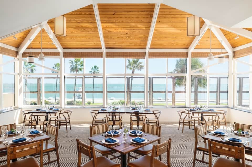 Beachfront dining room with floor-to-ceiling windows, ocean views, wooden ceiling beams, and dining tables