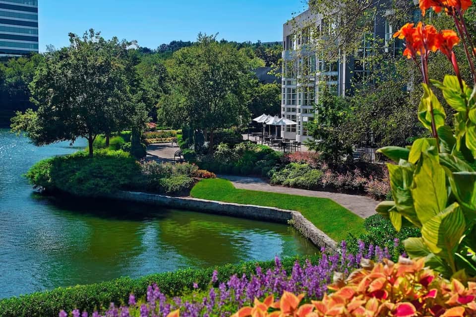 Waterfront hotel building overlooking river with lush trees, colorful flower gardens, and green manicured landscape
