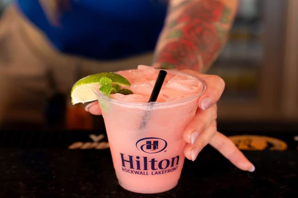 Pink cocktail with lime garnish labeled Hilton Rockwall Lakefront, held by person with colorful tattoo