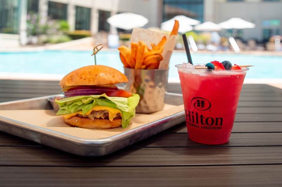 Burger, fries, and red cocktail on poolside table with Hilton branding and water view