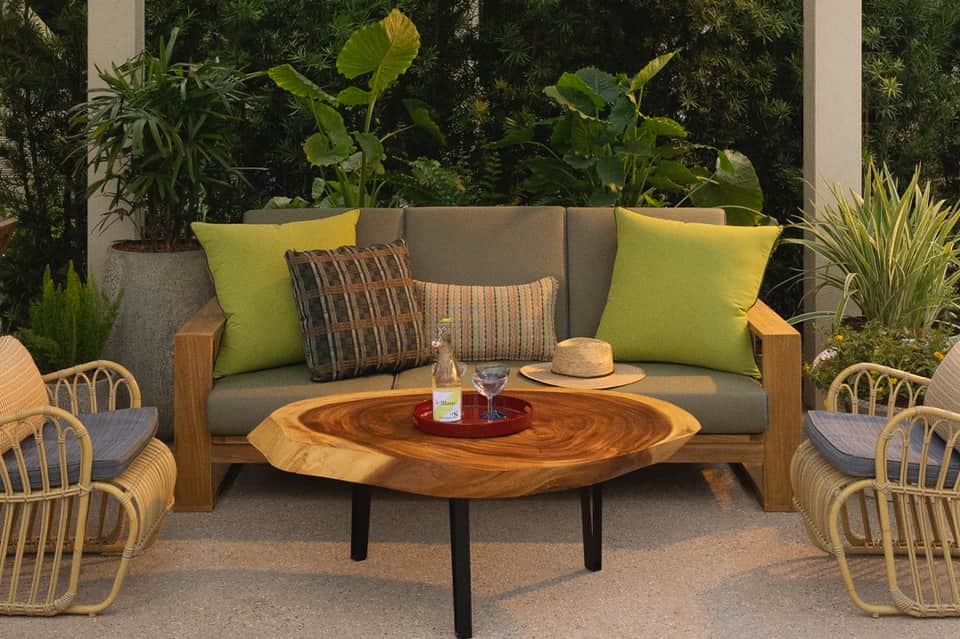 Outdoor patio seating with gray upholstered sofa, yellow pillows, wooden coffee table, rattan chairs, and lush garden backdrop